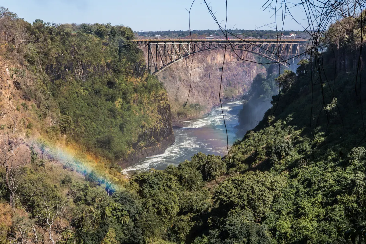 Un ponte ad arco in acciaio su una profonda gola verde con un fiume sottostante e un tenue arcobaleno nella spruzzi (il Ponte delle Cascate Vittoria, non il teatro)